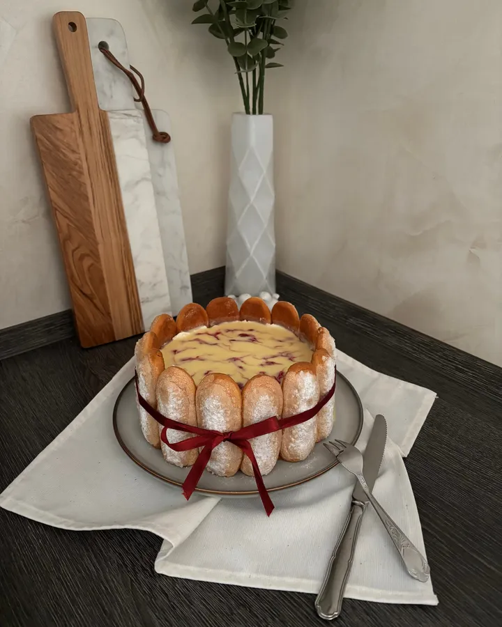 Charlotte ceinturée de biscuits à la cuillère et nouée d'un ruban rouge, le dessus marbré de coulis, sur une assiette posée sur un linge blanc.